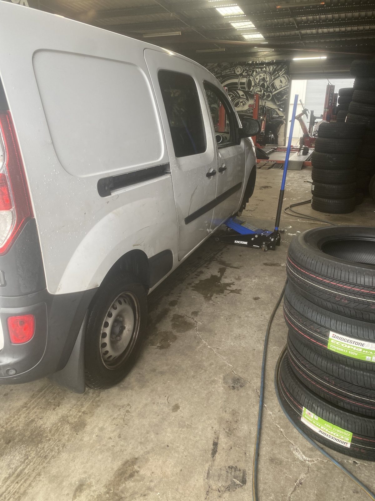 Silver van jacked up in the workshop bay beside a stack of new Bridgestone tyres